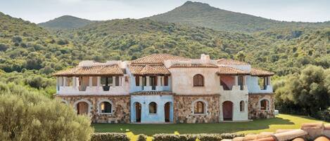 A large, multi-story building with a stone facade and tiled roof, surrounded by lush greenery and hills.
