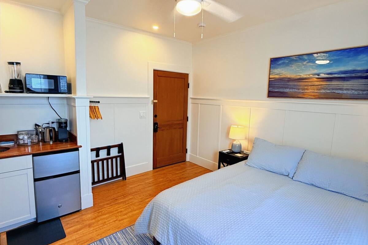 A bedroom with a bed, a desk, a chair, a nightstand, a lamp, a ceiling fan, and a painting on the wall.