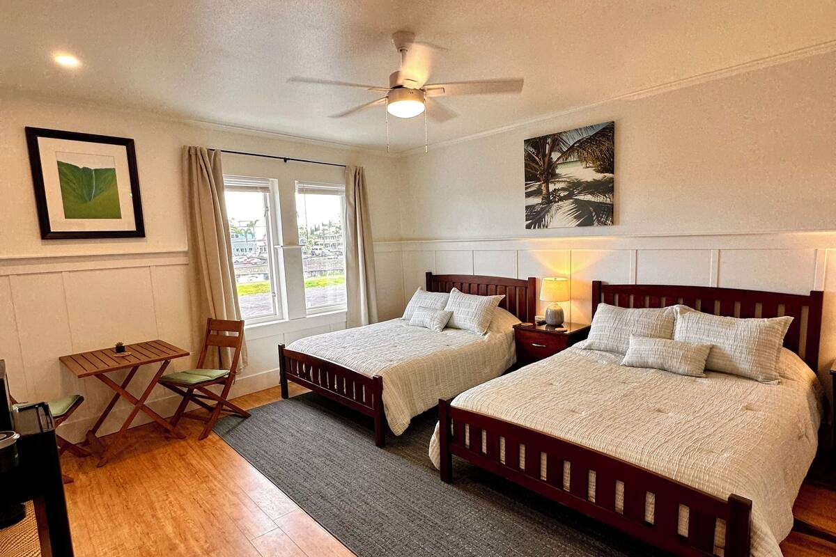 A bedroom with two beds, a ceiling fan, a window with curtains, and a small table with a chair.