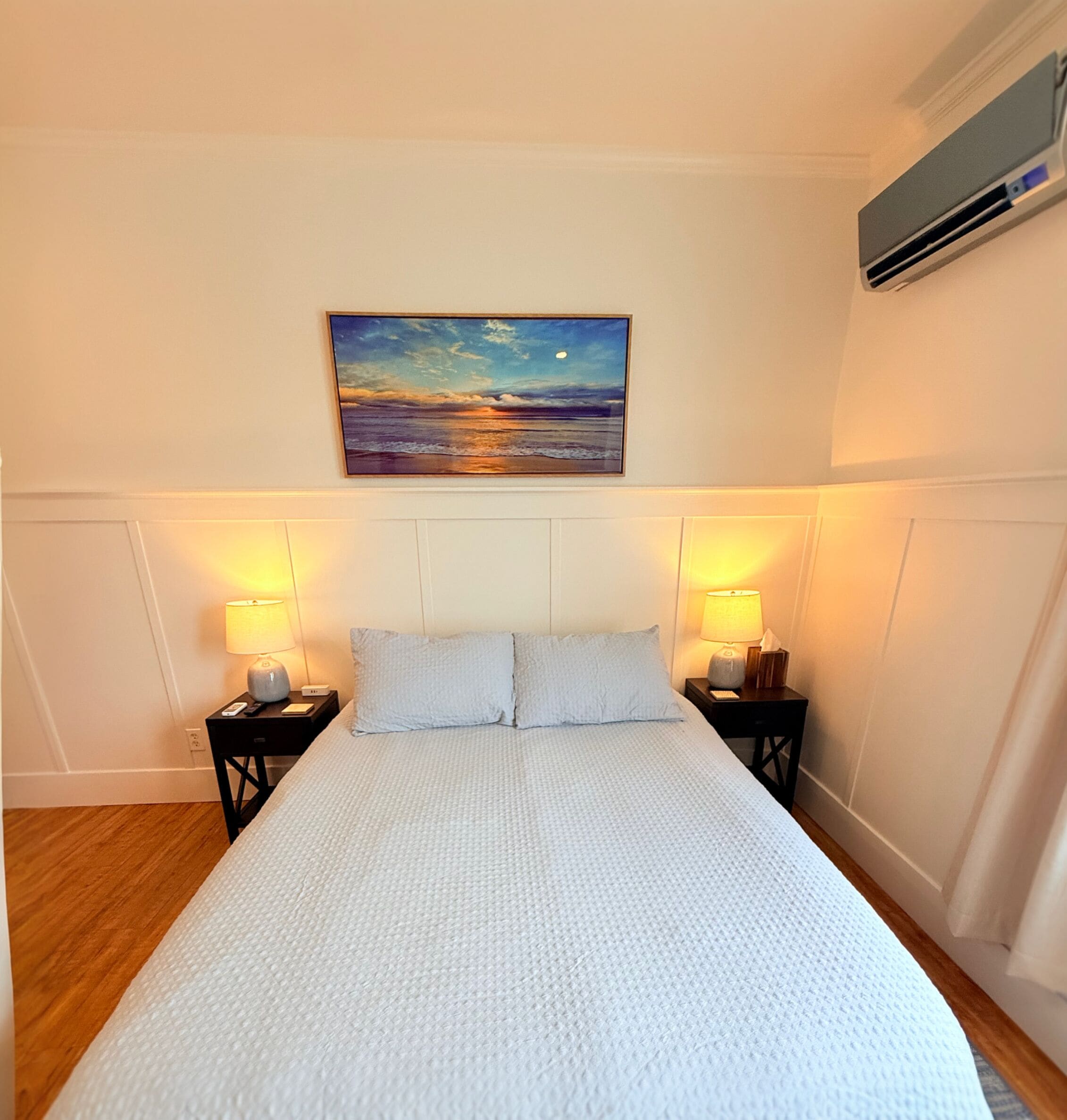 A hotel room with a large bed, two bedside tables with lamps, a painting of a sunset, and an air conditioning unit on the wall.