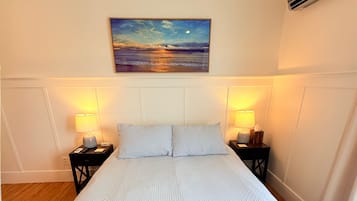 A hotel room with a large bed, two bedside tables with lamps, a painting of a sunset, and an air conditioning unit on the wall.