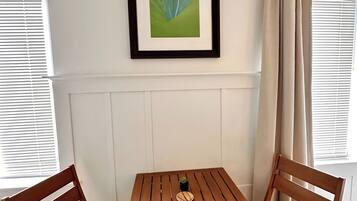 A corner room with a wooden table and two chairs, a framed picture on the wall, and a window with curtains.