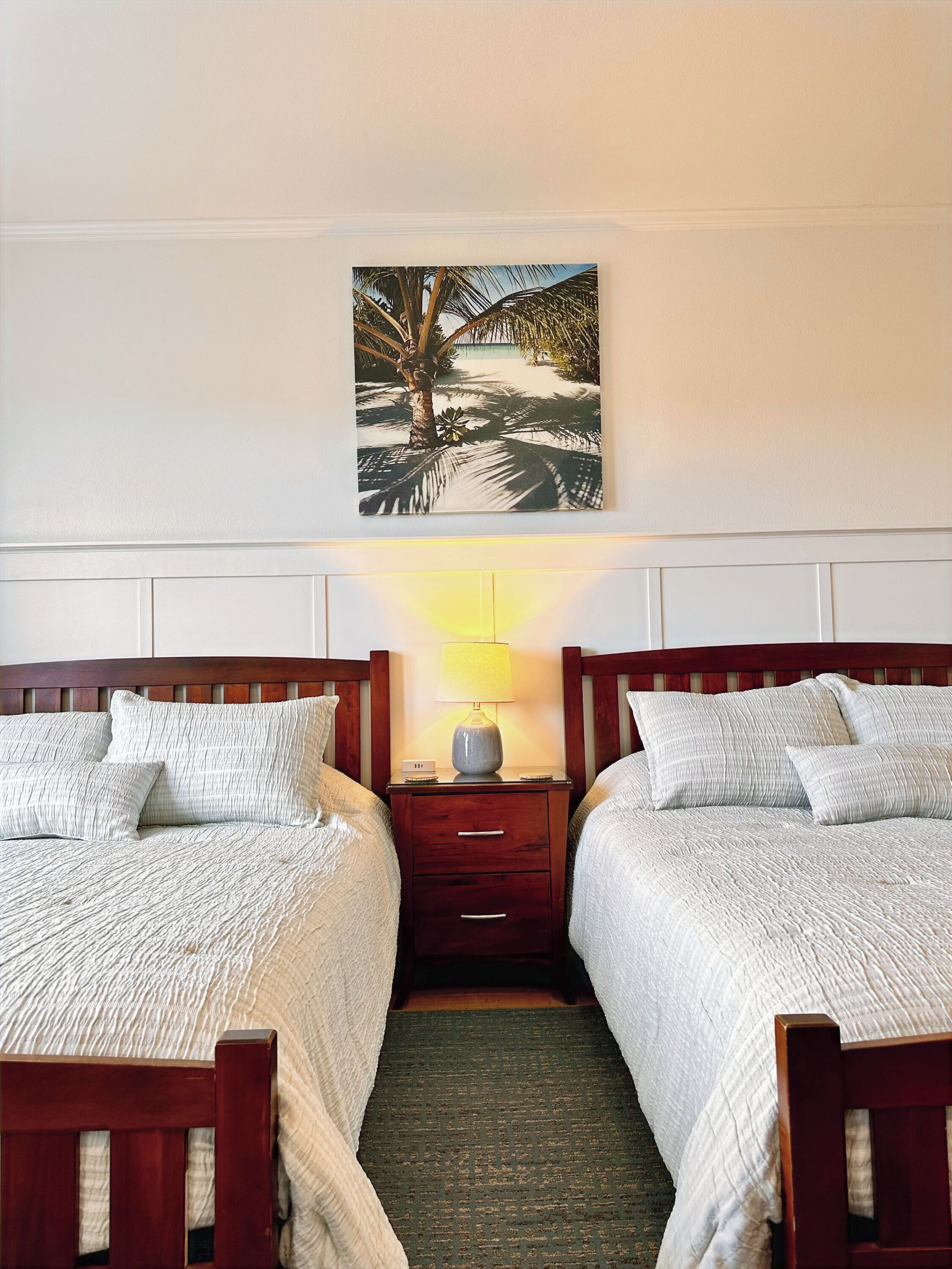 Ein Hotelzimmer mit zwei Einzelbetten, einem Nachttisch mit Lampe und einem Wandbild mit tropischer Szene.