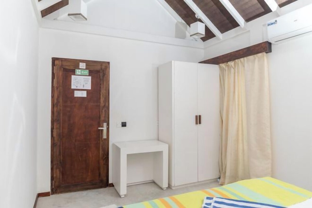 Una habitación pequeña con una puerta de madera, un armario blanco, un escritorio blanco y una cama con colcha a rayas.