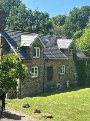 A stone building with multiple windows and a sloped roof, surrounded by a grassy area and trees.
