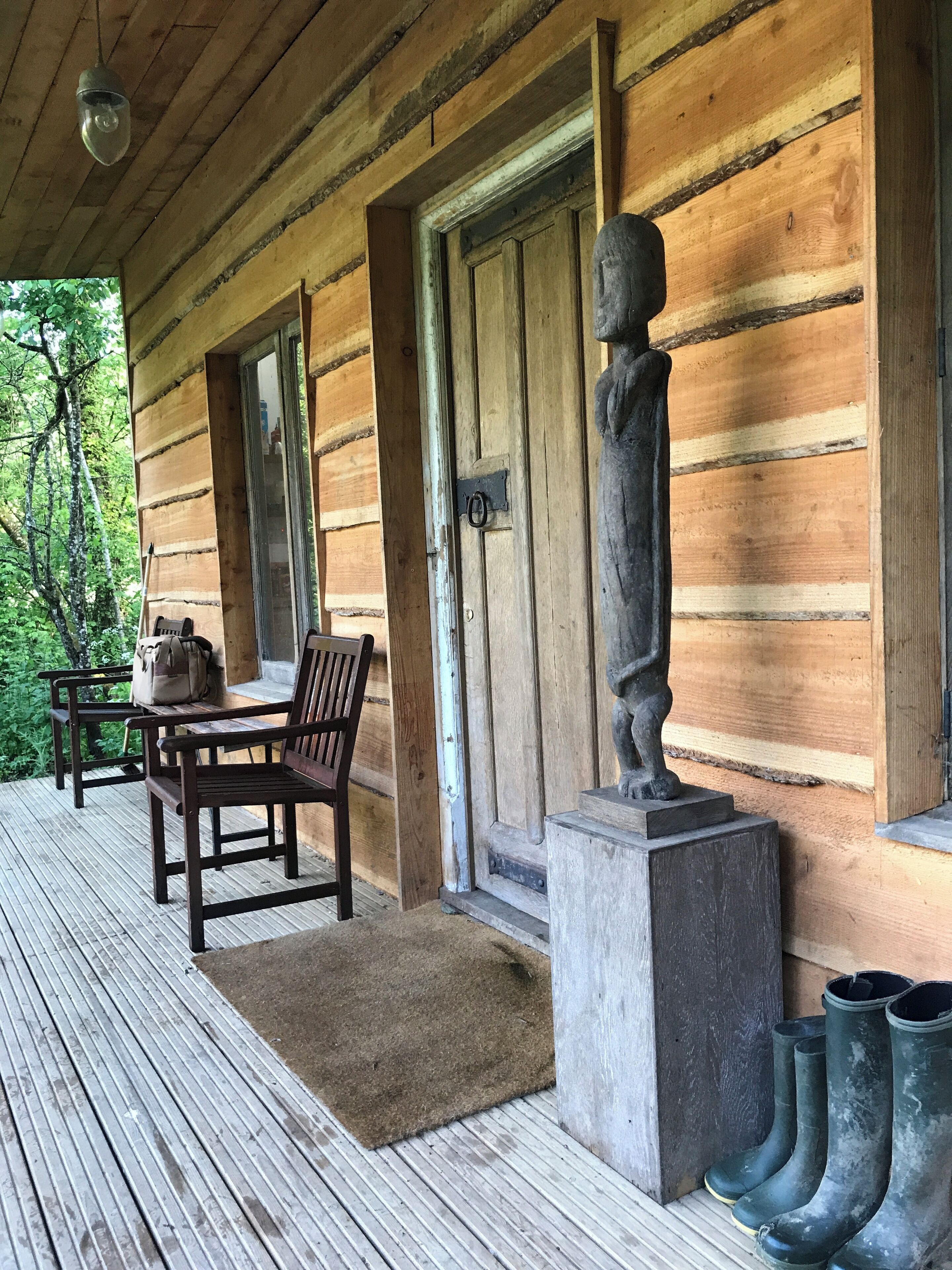 A wooden cabin with a porch, a bench, a table, and a statue.