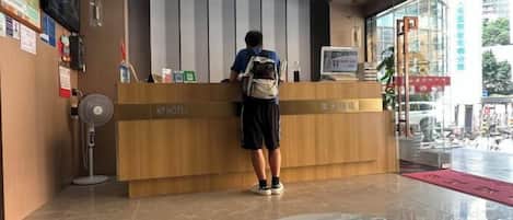 A hotel reception desk with a person standing behind it, a fan, and a glass partition.