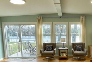 A room with a large sliding glass door leading to a deck with outdoor furniture. There are two armchairs with cushions and a small table with a lamp. - Cozy Riverfront Cabin: Parks,Kayaks& Island Charm (Momence)