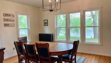 A dining room with a round wooden table and six chairs, large windows with a view of greenery, and a black chandelier.