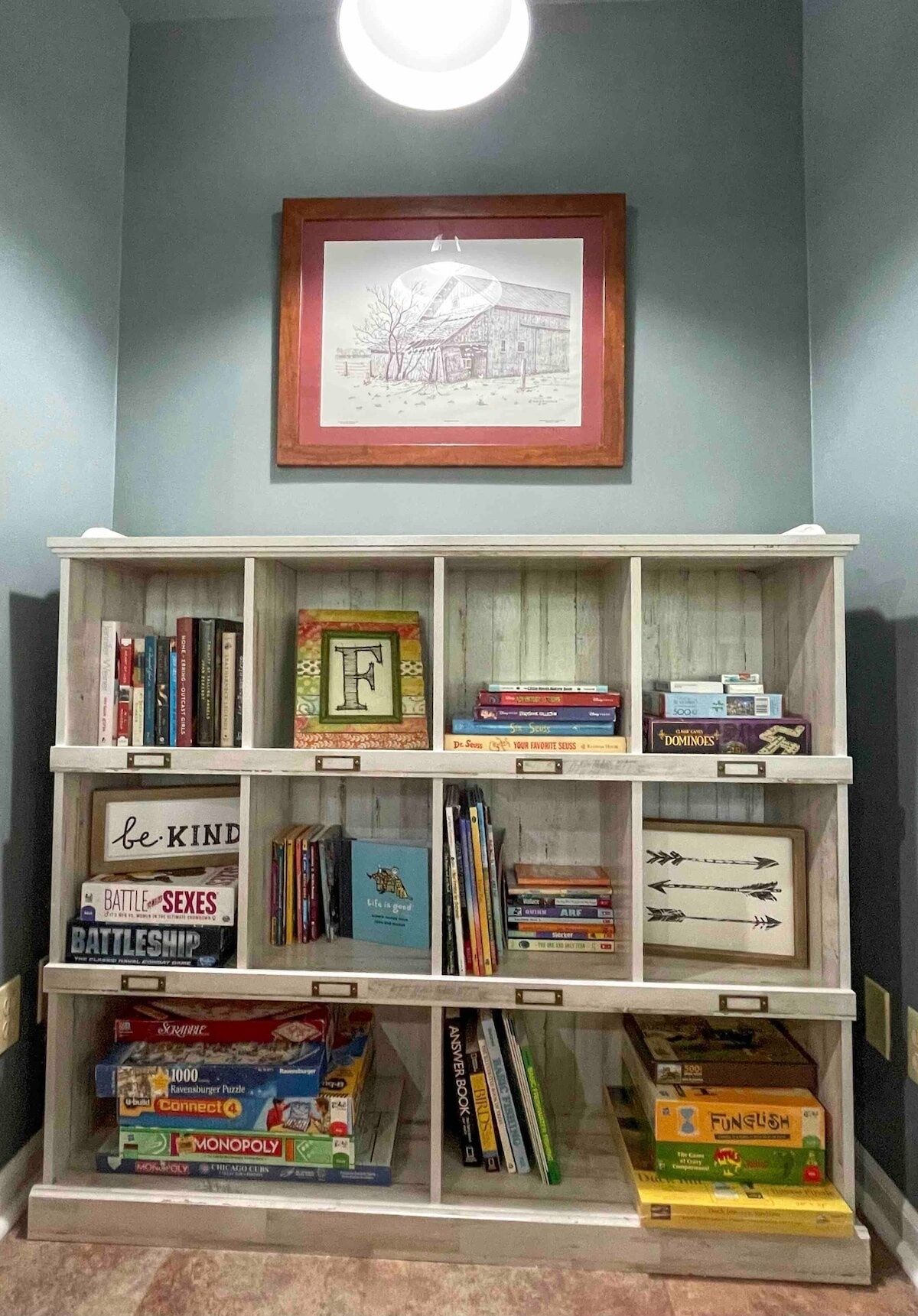 Uma estante com vários livros, jogos e um quadro na parede.