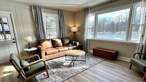 A living room with a brown leather sofa, two armchairs, a coffee table, and a lamp. There are large windows with curtains and a view of trees. - Escape to The Riverfront Hideaway (Momence)