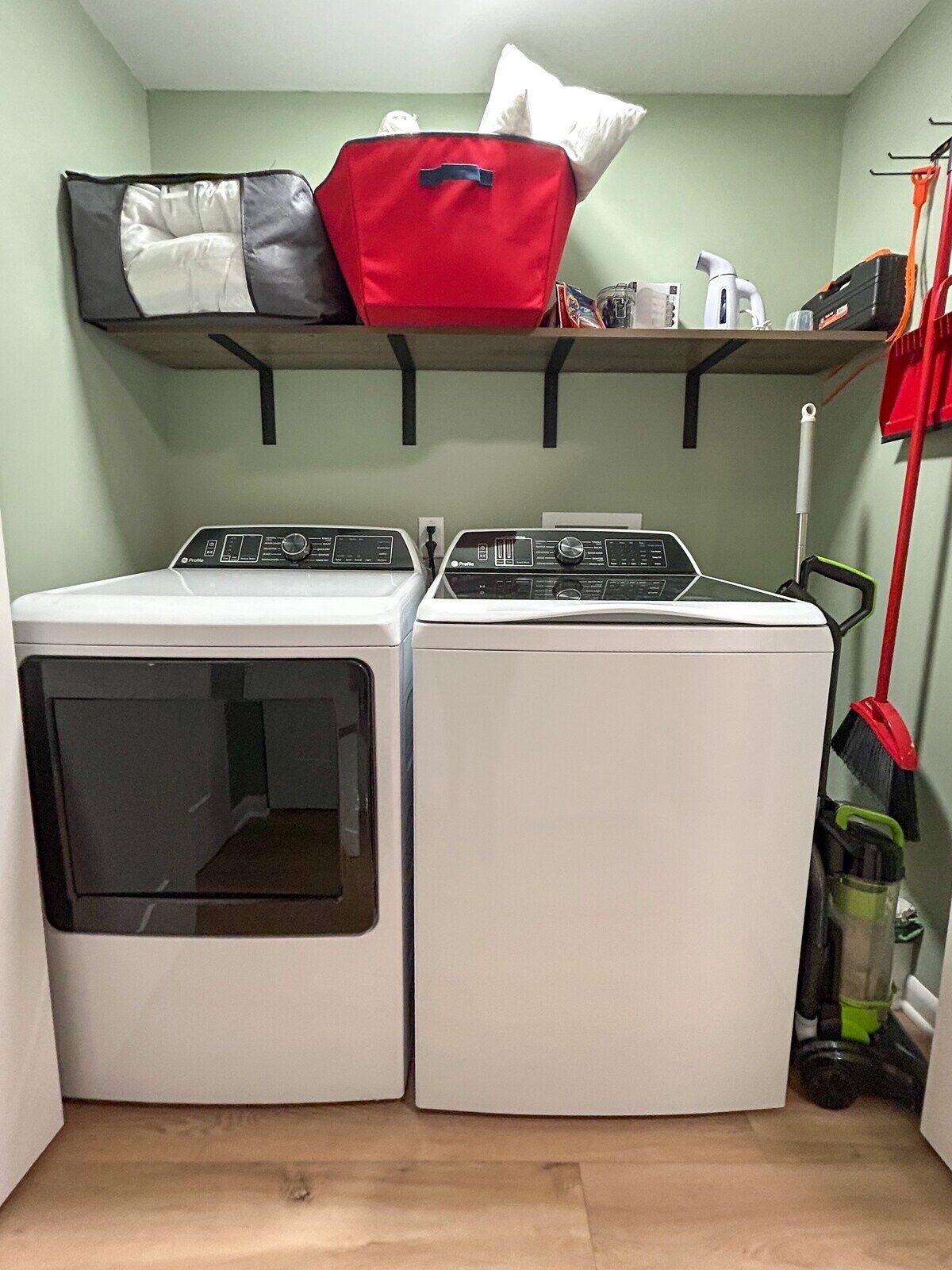 A laundry room with two stacked washing machines, a shelf with a red bag and pillows, and a broom leaning against the wall.