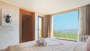 Une chambre d’hôtel avec une grande fenêtre, un lit recouvert d’une couette blanche, une table de chevet et une vue sur un paysage verdoyant.