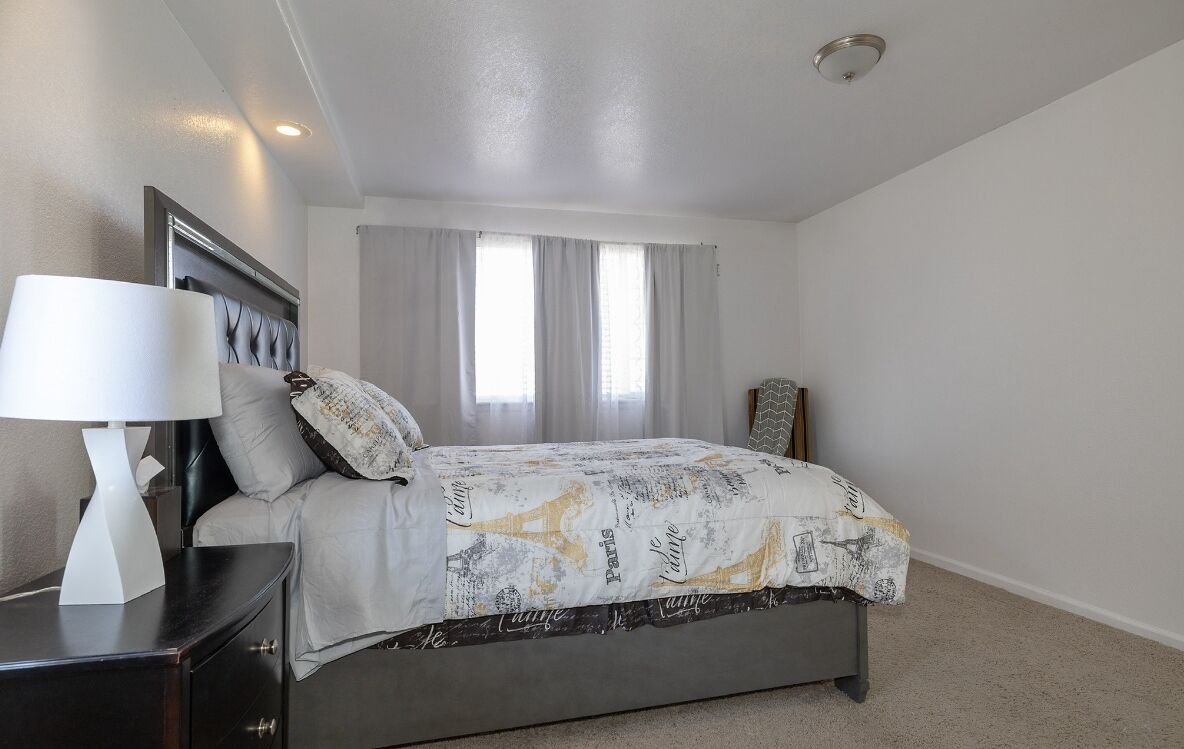 Une chambre à coucher comprenant un lit, une table de chevet, une lampe, une fenêtre avec des rideaux et un luminaire au plafond.