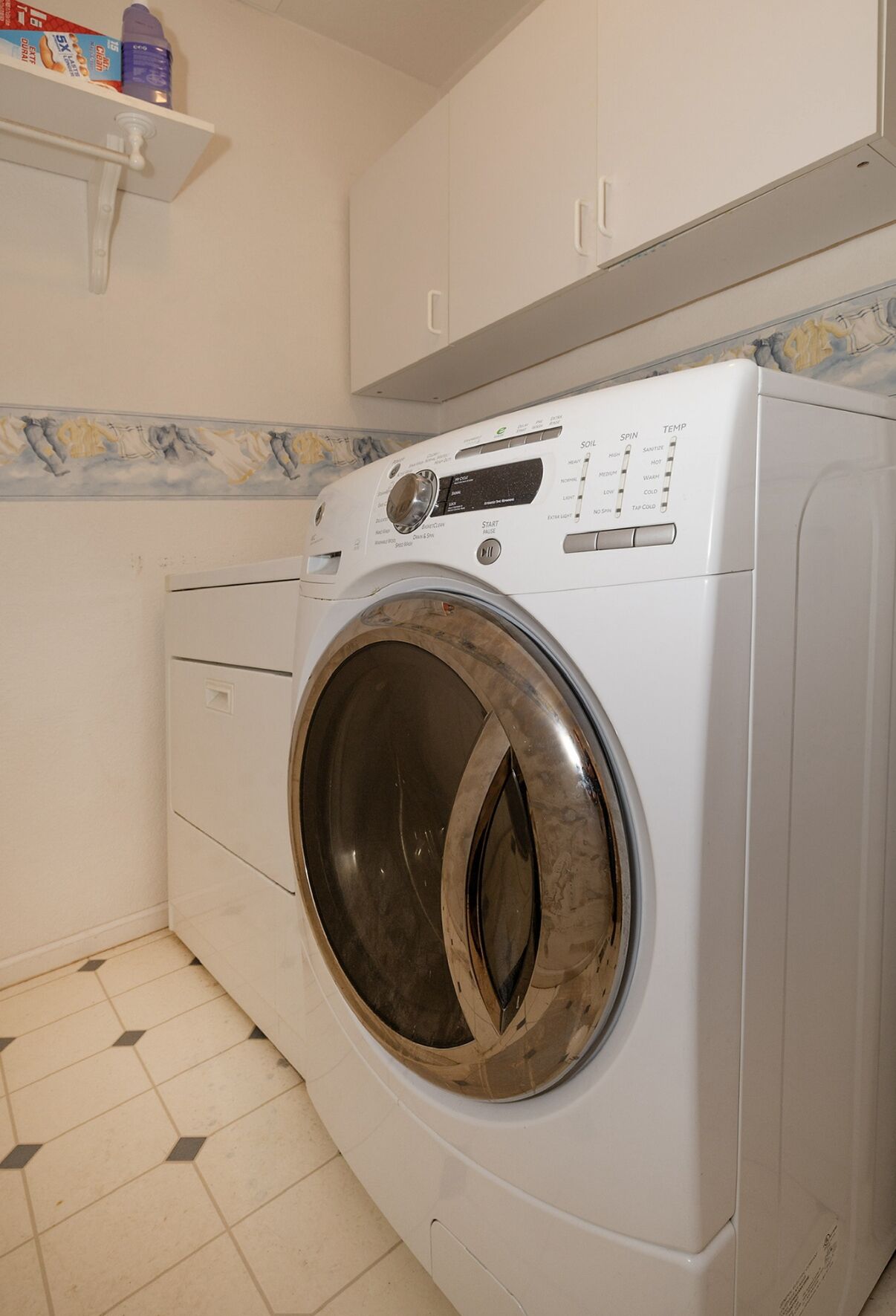 Una lavadora de carga frontal en un cuarto de lavado con piso de baldosas y un estante arriba con productos de limpieza.