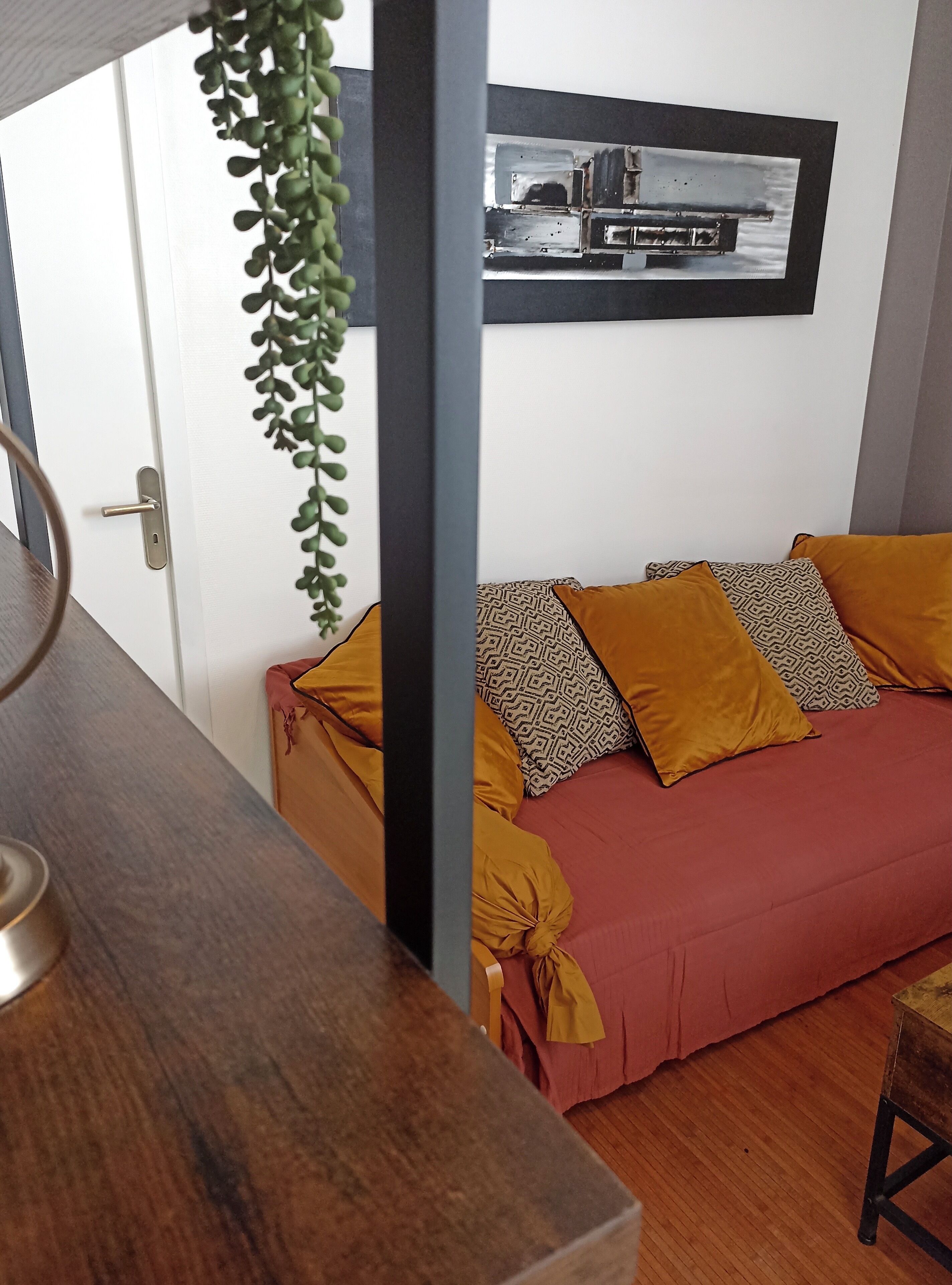 A room with a wooden table, a sofa with patterned cushions, a hanging plant, and a framed picture on the wall.