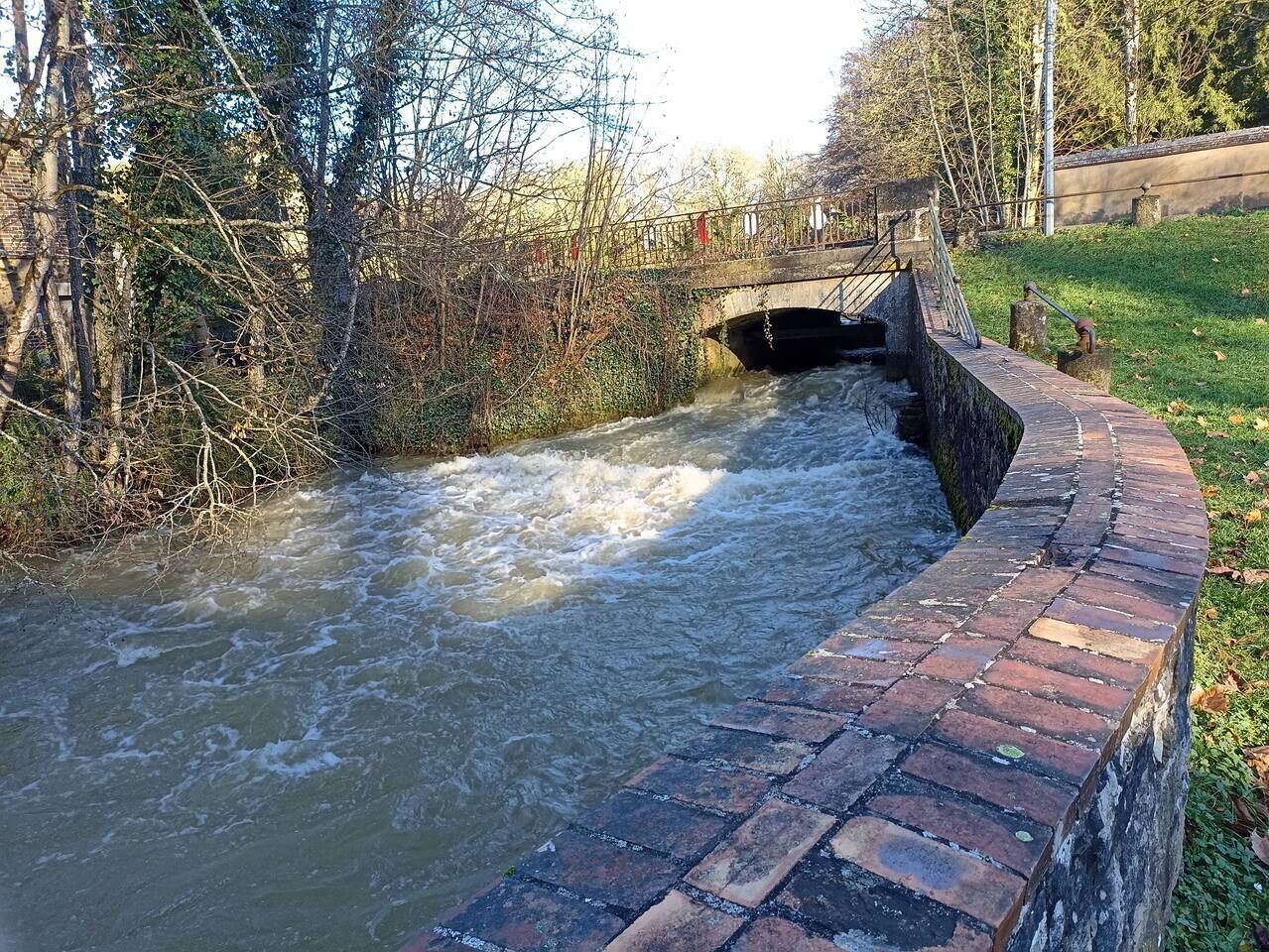 Un canal con paredes de ladrillo, un puente y árboles al fondo.