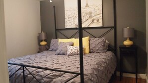 A bedroom with a bed, two bedside tables, a ceiling fan, and a large cityscape painting on the wall.