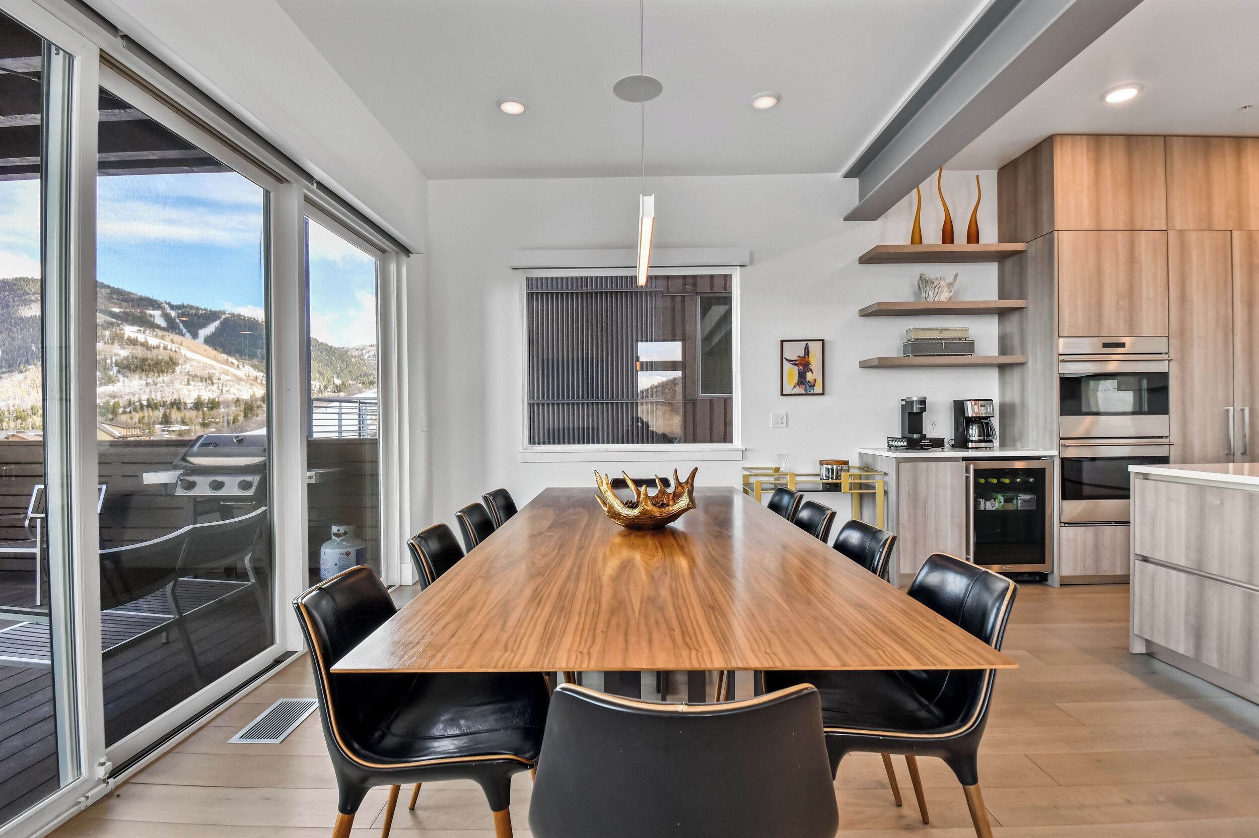 A modern kitchen with a wooden dining table, black chairs, and a view of mountains through large windows.