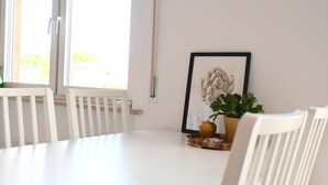 Une table à manger blanche avec des chaises, une œuvre d’art encadrée et une plante en pot, le tout placé devant un mur blanc.