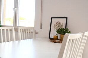 A white dining table with chairs, a framed artwork, and a potted plant on a white wall.