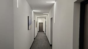 A long, narrow hallway with white walls and dark flooring, featuring recessed lighting and framed artwork on the walls.