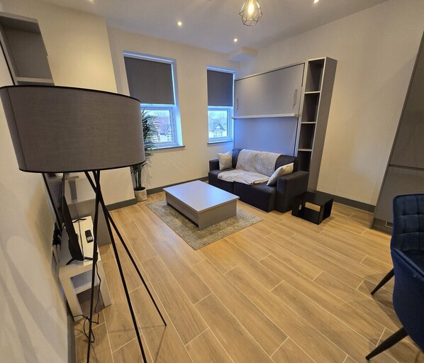 Een moderne woonkamer met een grijze bank, een salontafel en een tv-meubel. Twee ramen zorgen voor natuurlijk licht en uitzicht.