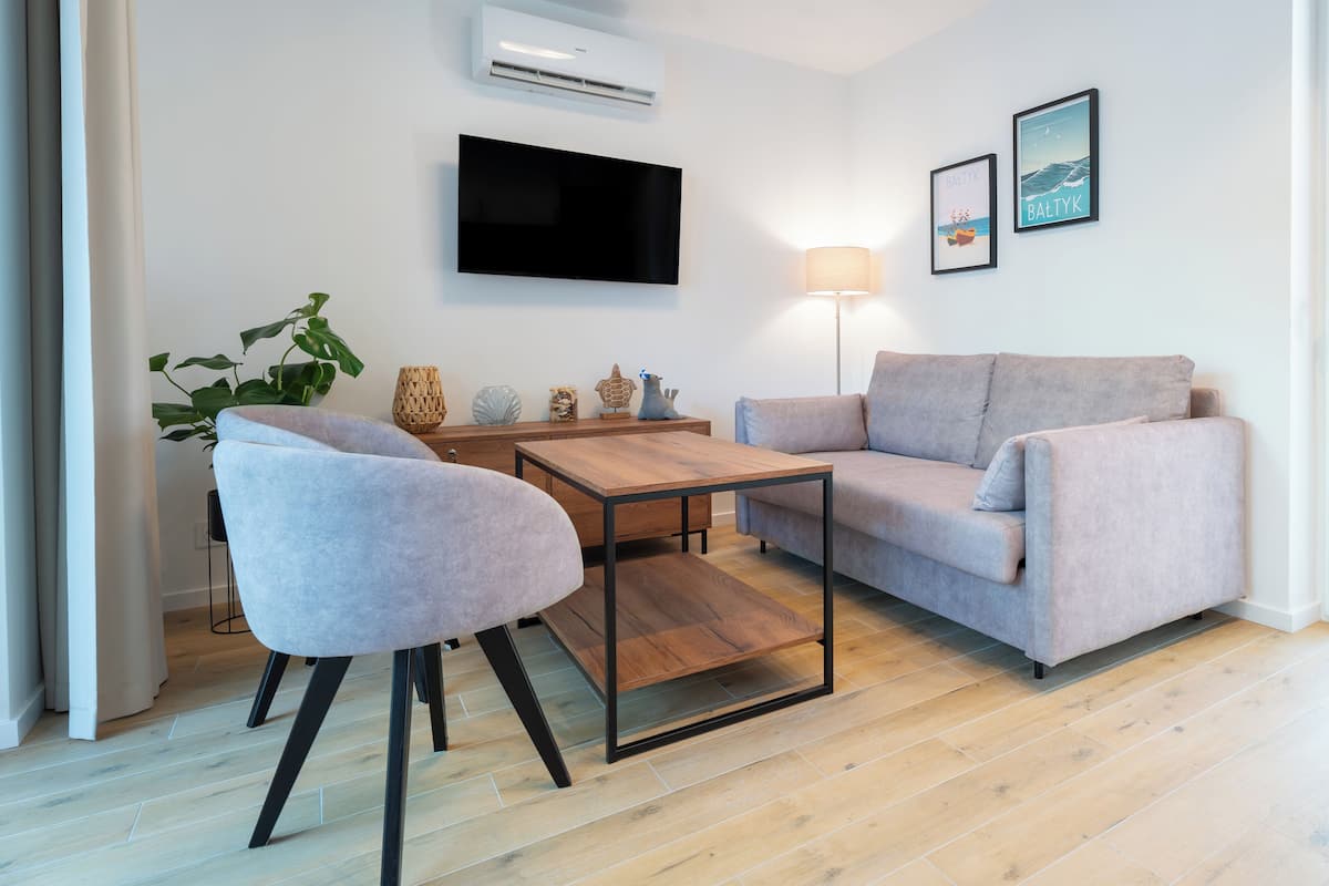 Una sala de estar moderna con un sofá gris, una mesa de centro de madera y un sillón gris. Hay un televisor de pantalla plana montado en la pared, una lámpara de pie y dos cuadros enmarcados.