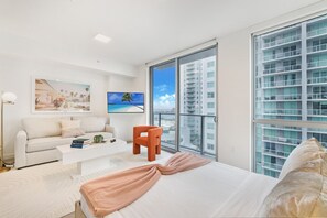 A modern bedroom with a large bed, a white sofa, a small orange chair, and a TV on the wall.
