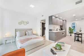 A modern bedroom with a large bed, bedside tables, and a nightstand. Adjacent to the bedroom is a kitchen with a countertop, sink, and cabinets. There is a dining area with a table and chairs.