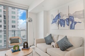 A modern living room with a large window, a sofa with patterned cushions, a floor lamp, and a coffee table with books and a plant. - Chic Downtown Studio With Rooftop Pool and Gym (Miami)