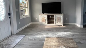 Un salon spacieux avec une grande fenêtre, un téléviseur à écran plat posé sur un meuble blanc et un tapis à motifs sur le sol.