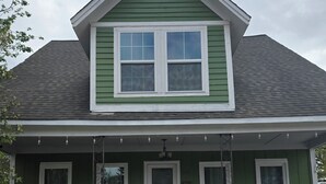 A green house with a front porch, two windows, a door, and a small porch area with chairs and a table.