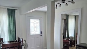 A spacious living room with hardwood flooring, a dining area with a wooden table and chairs, a large window with a decorative glass door, and a hallway leading to another room.