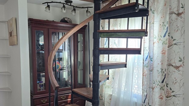 A wooden spiral staircase with a dark finish, leading to an upper floor. A large wooden cabinet with glass doors is visible in the background, and a patterned carpet covers the floor.