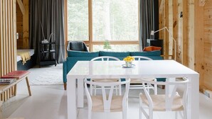 A wooden cabin with a loft bedroom, a dining area with white furniture, and a view of the forest through large windows.