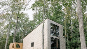 A modern wooden cabin with a large window and a smaller arched window, surrounded by a forest.