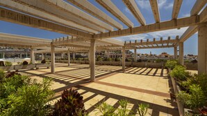 Een overdekt terras buiten, met een betegelde vloer, een houten pergola en potplanten.