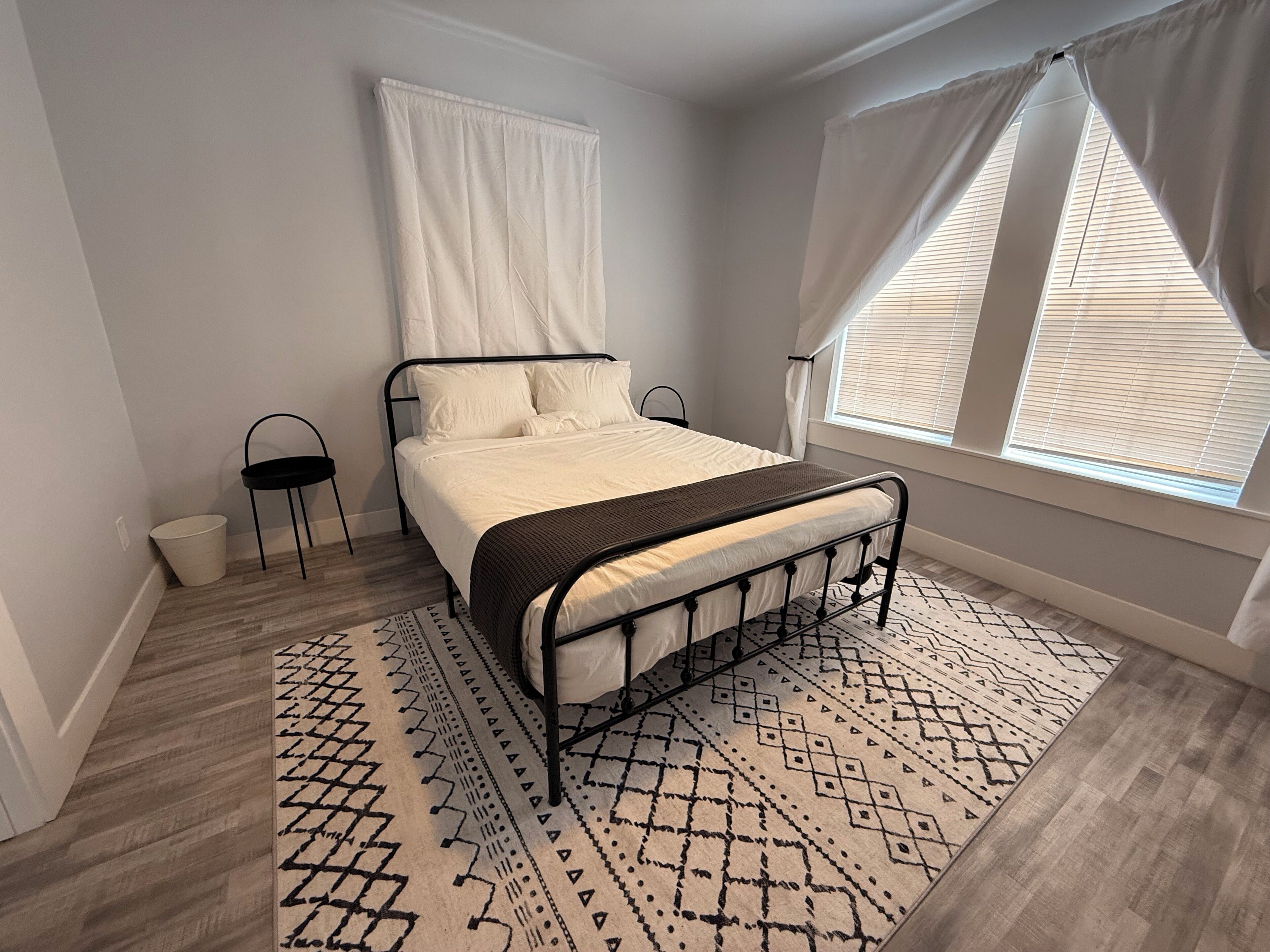 A bedroom with a bed, a window with blinds, a patterned rug, and a small table with a chair.