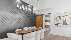 Un comedor moderno con una mesa de madera y sillas blancas tapizadas, un diseño geométrico en la pared y una lámpara colgante.