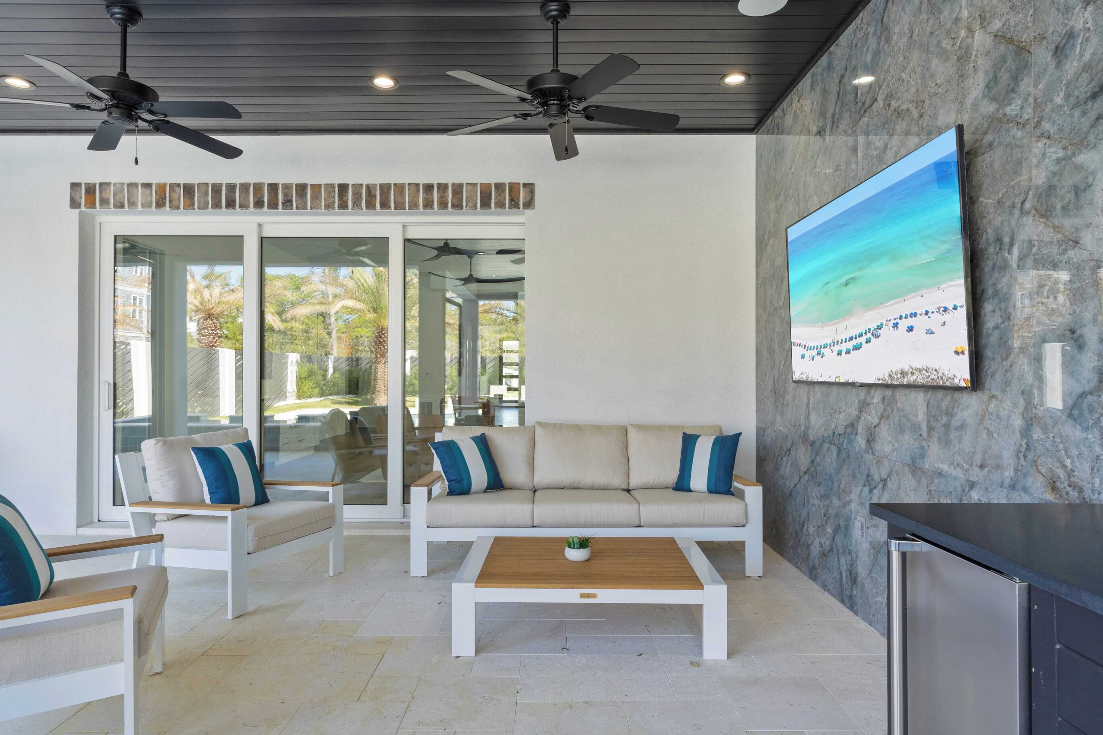 A modern outdoor living area with a sofa, chairs, a coffee table, ceiling fans, and a large beach painting on the wall.