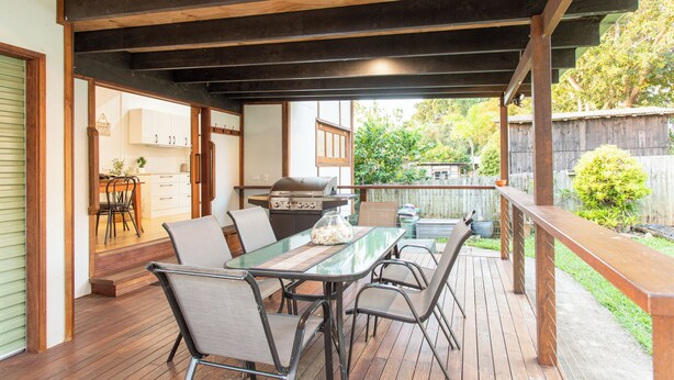 En udendørs terrasse med et bord med glasplade, metalstole og en grill.