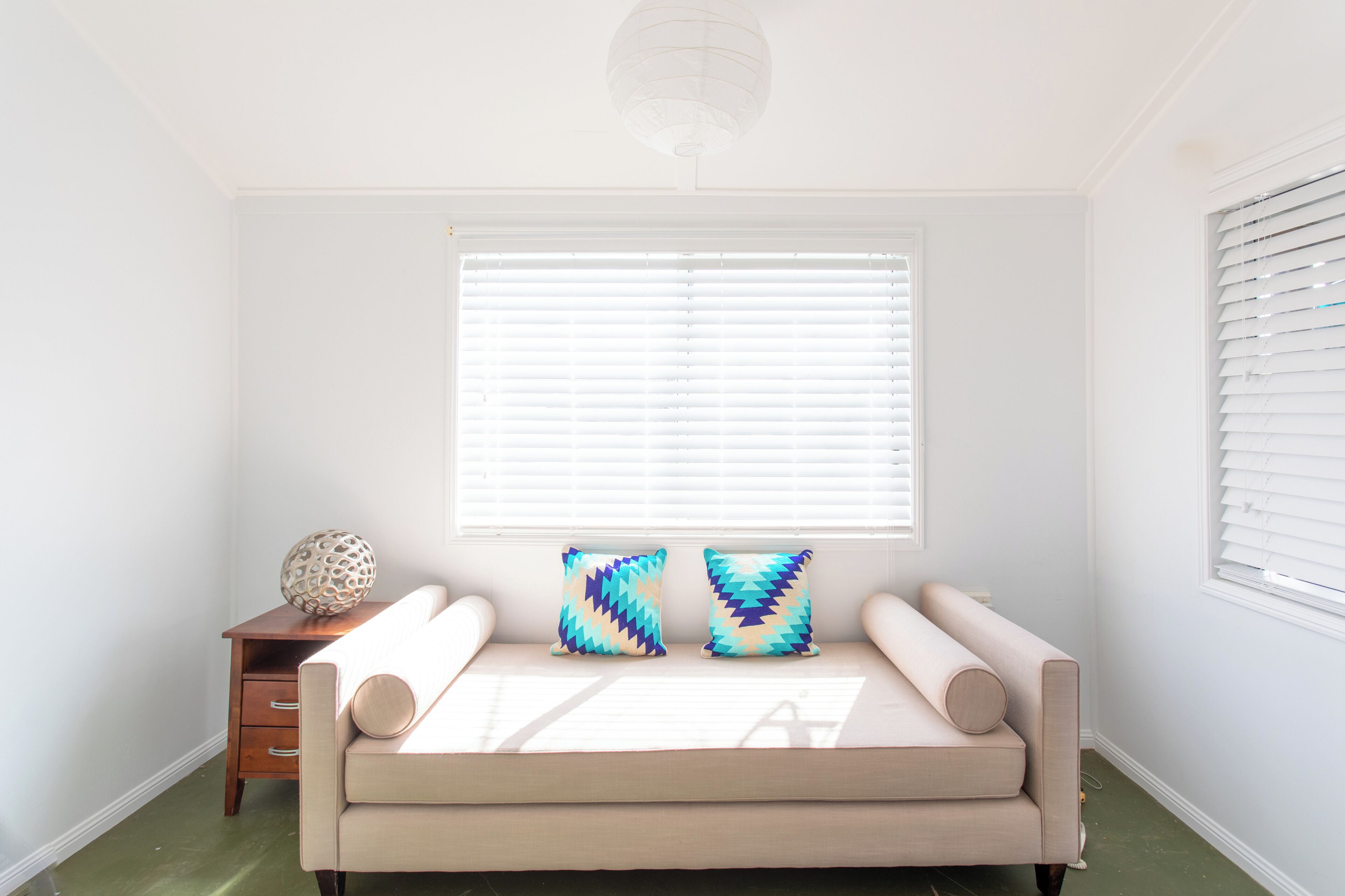 A room with a beige sofa, two patterned pillows, a wooden side table with a decorative sphere, and a large window with blinds.