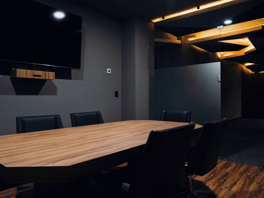 Una sala de conferencias con una gran mesa de madera, sillas negras, un televisor de pantalla plana montado en la pared e iluminación empotrada.
