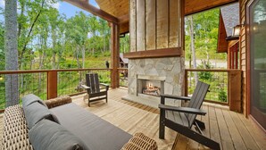 A wooden deck with a stone fireplace, outdoor seating, and a view of the forest.