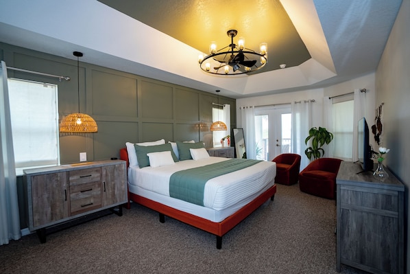 A bedroom with a large bed, a wooden dresser, a red chair, a plant, and a ceiling fan.