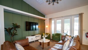 A living room with a brown leather sofa, a white coffee table, a green armchair, and a flat-screen TV mounted on the wall. - 2 King 2 Twin 1 Queen + Enormous Deck 3 BD Lakeview by Waterfront Park & Events (Clermont)