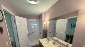 A bathroom with a double sink, a large mirror, a clock, and a shower with a grab bar.
