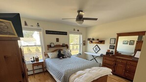 A bedroom with a bed, a dresser, a mirror, a ceiling fan, and a window with a view.