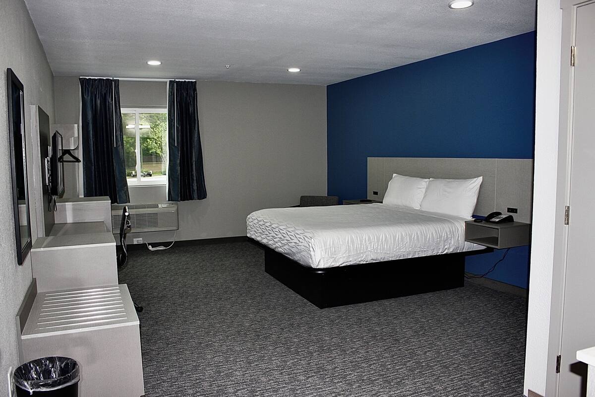 A hotel room with a bed, a desk, a chair, a trash bin, a window with curtains, and a wall-mounted heater.